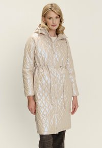 Donna che indossa un cappotto con cappuccio trapuntato beige chiaro, con coulisse e chiusure a scatto, abbinato a pantaloni a quadri scuri.