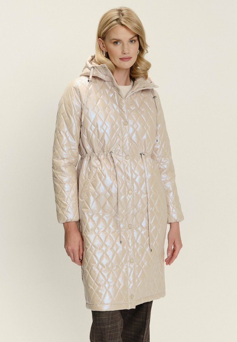 Donna che indossa un cappotto con cappuccio trapuntato beige chiaro, con coulisse e chiusure a scatto, abbinato a pantaloni a quadri scuri.