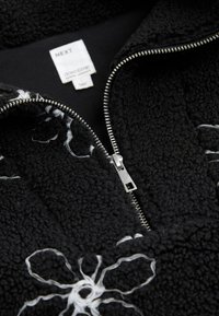 Sort fleecejakke med hvid blomstbroderi, der har en sølvlynlås og en mærkat med brandnavnet "NEXT" og produktdetaljer.