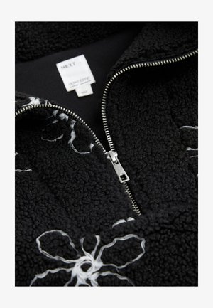 Veste polaire noire avec broderie de fleurs blanches, dotée d'une fermeture éclair argentée et d'une étiquette indiquant le nom de la marque « NEXT » ainsi que les détails du produit.