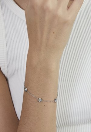 Silver chain bracelet with small flower-shaped charms worn on a wrist against a white ribbed fabric background.