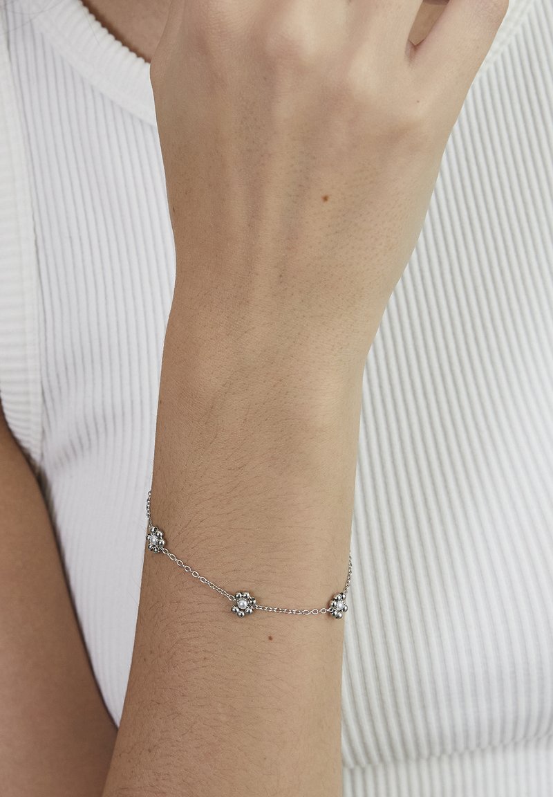 Silver chain bracelet with small flower-shaped charms worn on a wrist against a white ribbed fabric background.