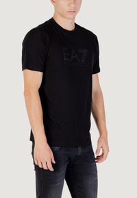 T-shirt in cotone nero con colletto rotondo e maniche corte. Presenta il logo EA7 impresso sul davanti. Indossato con jeans in denim scuro.