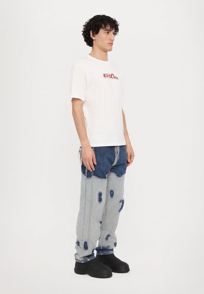 T-shirt blanche à manches courtes avec impression graphique rouge sur la poitrine, associée à un jean en denim bleu et gris déchiré, et des chaussures noires massives.