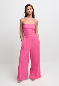 Fuchsia stropløs jumpsuit med en rynket overdel, vide ben og sidelommer. Glat stof med en behagelig pasform. Teksturerede beige sko.