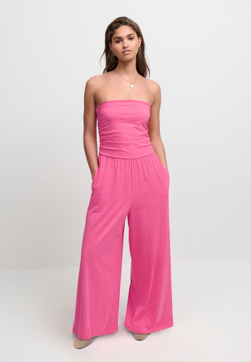 Fuchsia stropløs jumpsuit med en rynket overdel, vide ben og sidelommer. Glat stof med en behagelig pasform. Teksturerede beige sko.