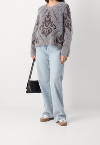 Osoba oblečená v šedém vzorovaném huňatém svetru, světle modrých širokých džínech, bílých teniskách s béžovými podrážkami, držící černou kabelku.