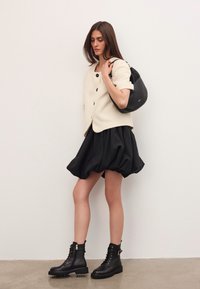 Femeie îmbrăcată cu o jachetă crem cu mâneci scurte �și nasturi, fustă bubă neagră, bocanci negri cu șireturi și ținând o geantă de umăr din piele neagră.