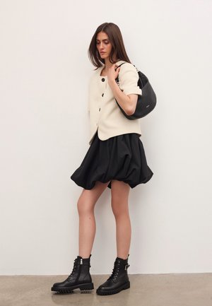 Sieviete, kas valkā krēmkrāsas īsām piedurknēm pogātu jaku, melnu burbuļsvārku, melnus šņorējamus zābakus un tur rokās melnu ādas plecu somu.