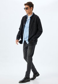 Chaqueta de mezclilla negra sobre una camisa de botones azul claro, combinada con jeans gris oscuro y botines negros. Lleva gafas de sol.
