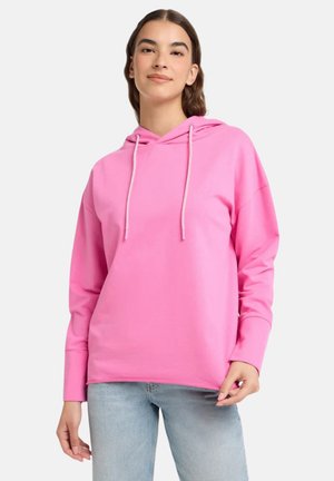 Woman wearing a bright pink hoodie with drawstrings and light blue jeans, standing against a plain light background.