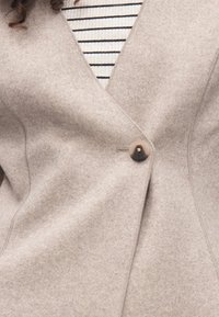 Lichtgrijs blazer van een wolmix met een V-hals ontwerp en een enkele bruine knoop, met subtiele stiksels en een gladde textuur. Gestreepte top eronder.