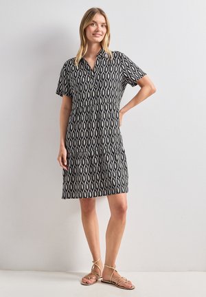 Femme debout, main sur la hanche, portant une robe courte à manches courtes, noire et blanche à motifs, et des sandales plates beiges avec des brides.