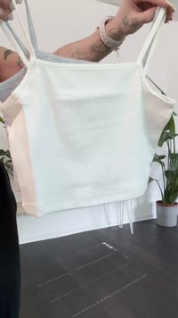 Osoba trzymająca białą bluzkę na ramiączkach na jasnym tle, w tle widać zielone rośliny i doniczkę z rośliną.
