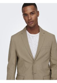 Blazer de lino beige con dos botones, solapas de muesca y un bolsillo en el pecho. Llevado sobre una camiseta blanca de cuello redondo.