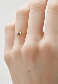 Anillo de oro con tres pequeñas piedras y una banda lisa. Llevado en una mano de color claro, mostrando intrincados detalles cerca de las piedras.