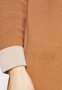 Gros plan sur le bras d'une personne portant un pull en maille marron avec un revers beige clair.