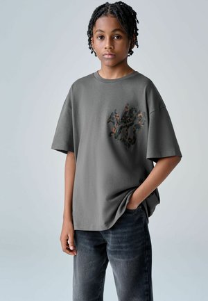 Graues übergroßes T-Shirt aus Baumwolle mit Tarnmuster und dunklem Logo. Kombiniert mit dunklen Denim-Jeans.