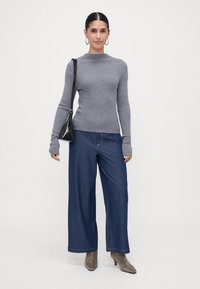 Moteris, aps wearing g Grey juostuota megztinis, plačiakelnės mėlynos kelnės, pilki aulinių batai, kabinamos auskarai ir nešanti juodą rankinę prieš baltą foną.