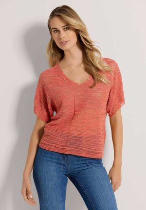 Vrouw met lang blond haar, gekleed in een losse koraalkleurige gebreide top met korte mouwen en een blauwe spijkerbroek, staande tegen een eenvoudige lichte achtergrond.