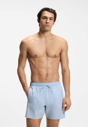 Junger Mann mit dunklen Haaren, der hellblaue Badeshorts trägt und mit einer Hand in der Tasche vor weißem Hintergrund steht.