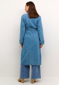 Kvinna vänd bort från kameran iklädd en lång blå denimkappa med skärp, vida denimbyxor och beige ankelboots med klack mot en enfärgad bakgrund.