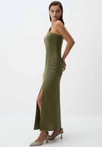 Olivgrünes, figurbetontes Maxikleid mit einseitigem Trägerdesign, seitlichem Schlitz und glatter Textur. Kombiniert mit hellfarbigen High Heels.