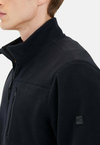 Schwarze Fleecejacke mit hohem Kragen, Reißverschluss-Brusttasche und strukturiertem Stoff. Verfügt über kontrastierende Nähte und ein kleines Logo am Arm.