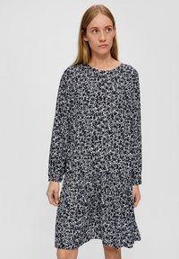 Frau mit langen blonden Haaren, die ein marineblaues Kleid mit weißem Blumenmuster trägt, steht vor einem schlichten grauen Hintergrund.