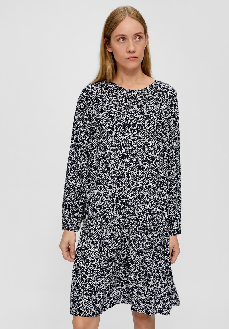 Frau mit langen blonden Haaren, die ein marineblaues Kleid mit weißem Blumenmuster trägt, steht vor einem schlichten grauen Hintergrund.