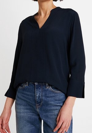 Femme portant un chemisier bleu marine foncé à manches longues avec un col en V, rentré dans un jean en denim bleu, debout devant un fond uni.