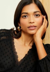 Blusa nera con texture a pois e rifiniture in pizzo, caratterizzata da un colletto a V e maniche a sbuffo. Orecchini a cerchio dorati completano il look.