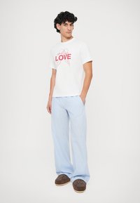 Witte T-shirt met rode "LOVE" graphic, gecombineerd met lichtblauwe gestreepte wijde broek en bruine instappers. Effen achtergrond.