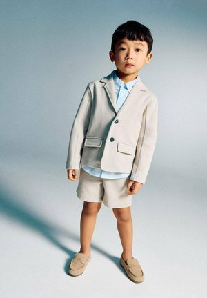 Băiat tânăr în picioare, purtând un sacou bej deschis, pantaloni scurți asortați, cămașă albastră cu guler și pantofi loafer bej, pe un fundal simplu gri.