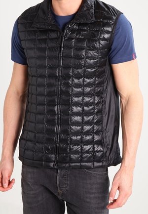 Homme portant un gilet sans manches matelassé noir sur une chemise bleue à manches courtes et un jean foncé, debout les bras détendus.