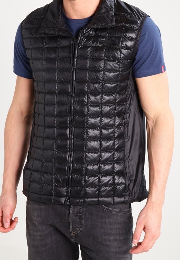 Homme portant un gilet sans manches matelassé noir sur une chemise bleue à manches courtes et un jean foncé, debout les bras détendus.