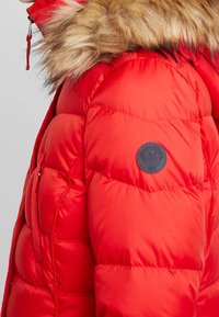 Rød pufferjakke med pelsforing i hætten og cirkulær sort logo-patch på venstre ærme.