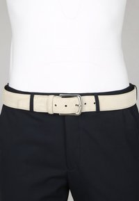 Beige suède riem met zilveren gesp om de taille, over zwarte broek en een witte ingestopte shirt.