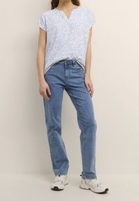 Person wearing light blue floral short-sleeve top, high-waisted blue jeans, and white sneakers standing against beige background.