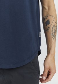 Námornícky modré tričko vyrobené z mäkkej textílie, s okrúhlym lemom, štítkom so slovom "BLEND" na boku a podrobným tetovaním na ruke.