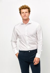 Camicia bianca a maniche lunghe con un design slim fit e chiusura a pulsante, abbinata a pantaloni blu scuro con un bottone frontale. Tessuto dalla consistenza liscia.