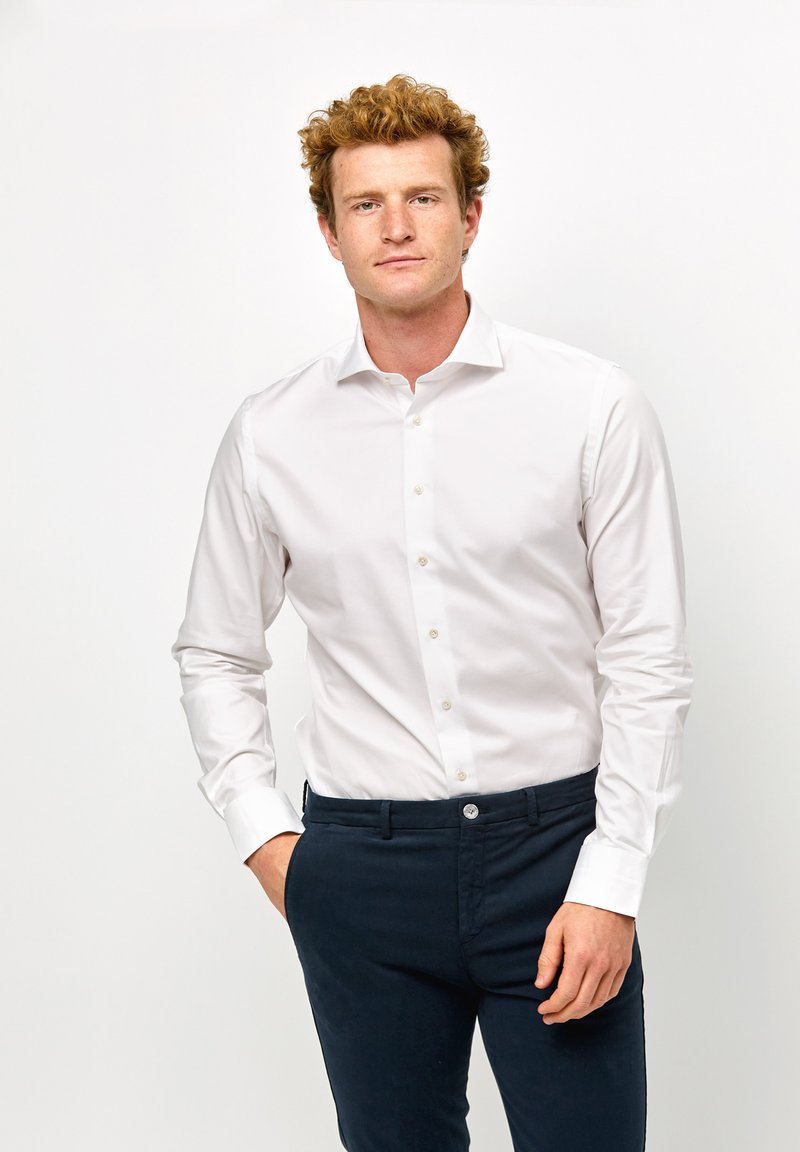 Camicia bianca a maniche lunghe con un design slim fit e chiusura a pulsante, abbinata a pantaloni blu scuro con un bottone frontale. Tessuto dalla consistenza liscia.
