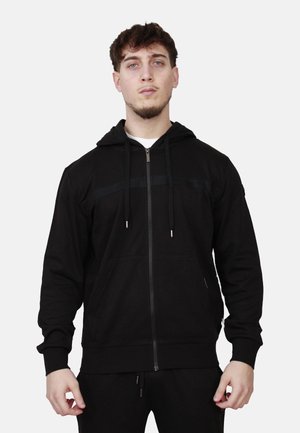 Giovane uomo che indossa una felpa nera con zip e coulisse, in piedi davanti a uno sfondo grigio chiaro uniforme, rivolto in avanti con espressione neutra.