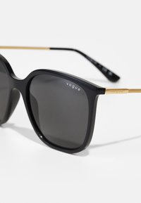 Melnie pārliekamie saulesbrilles ar tumšiem stikliem, plānām zelta metāla kājiņām un "vogue" zīmolu uz abiem stikliem un kājiņām. Gluda, spīdīga virsma.