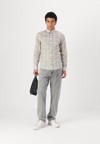 Lichtgepatroneerde overhemden met kraag, grijze broek en witte sneakers. Houdt een zwarte tas vast. Egaal achtergrond.