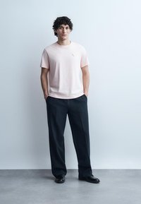 Hombre joven con cabello rizado, de pie con las manos en los bolsillos, viste una camiseta rosa claro, pantalones negros de pierna ancha y zapatos negros, sobre un fondo liso.