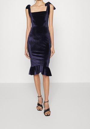 Robe midi sans manches en velours bleu marine avec encolure carrée, nœuds aux épaules, ourlet à volants, portée avec des sandales à talons noires à brides.