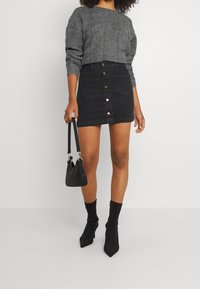 Pull en maille gris, mini-jupe en denim noir avec fermeture à boutons sur le devant, bottines noires et petit sac à main noir.