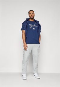 Camiseta de algodón navy con el texto "Yankees", pantalones cargo grises y zapatillas blancas. Atuendo casual con sudadera colgada sobre los hombros.
