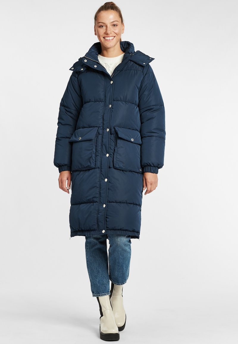 Navy blue padded winter coat with a hood, snap buttons, two front pockets, and ribbed cuffs, paired with light jeans and beige ankle boots.